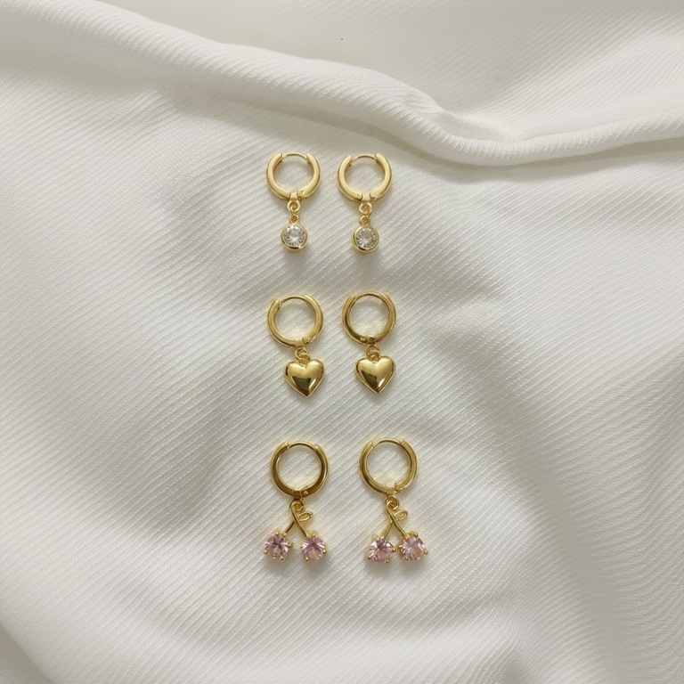 3 Set of Anti-Tarnish gold hoop earrings set includes one pairs with clear round stones, one pair with dangling gold hearts, and one pair with dangling gold cherub motifs adorned with small pink gemstones.