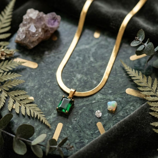 Gold necklace with a green gemstone pendant on a dark surface with leaves and crystals.