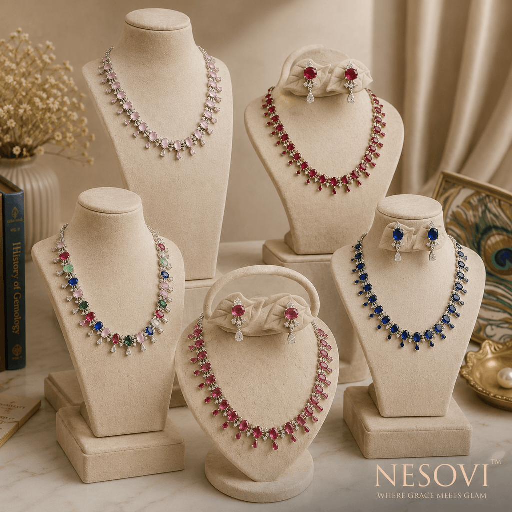 Five jewelry busts display necklaces, a bow, and matching earrings in a styled studio setting. The pieces feature silver-toned metal with clear stones and colored gemstone accents including pink, blue, green, and red.