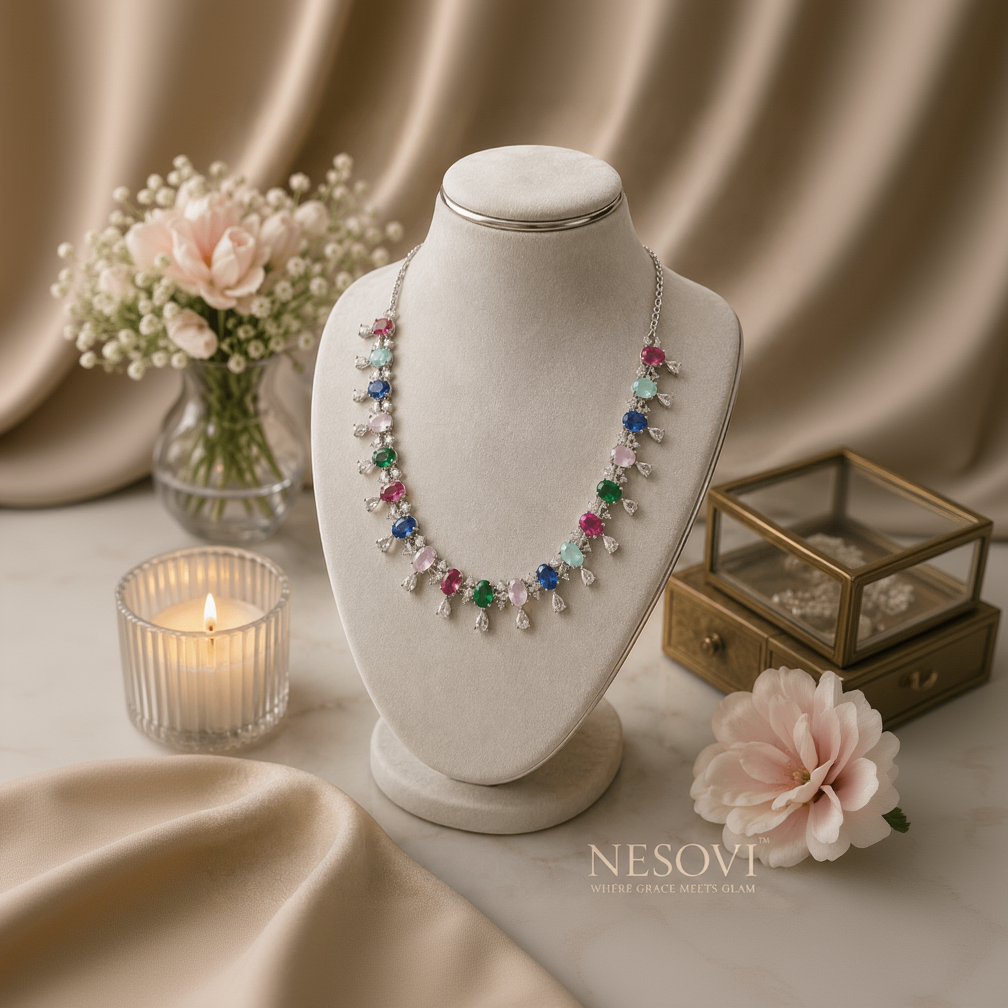 A colorful gemstone necklace displayed on a cream jewelry bust. The necklace features silver-toned metal with teardrop-shaped clear stones and vibrant multicolored gemstone accents in pink, blue, green, and light blue.